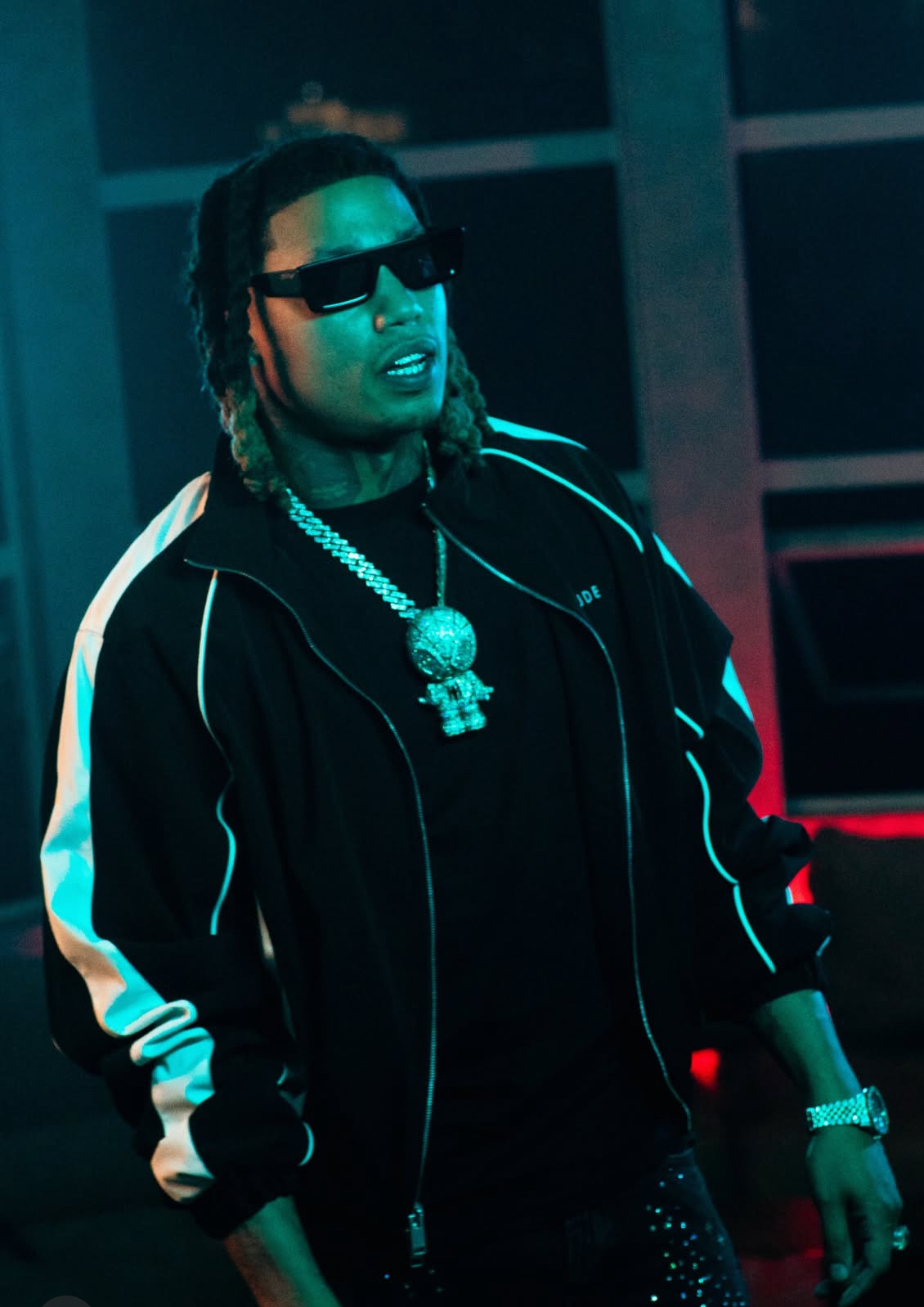Artist TEC in a striped track jacket and diamond chains under teal lighting